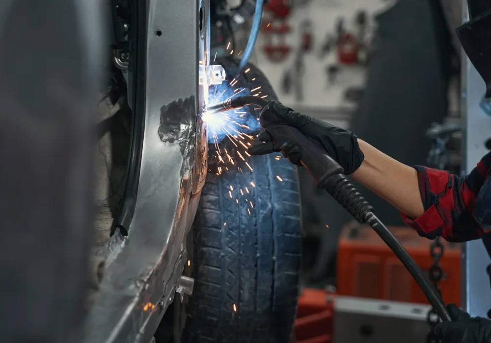 A mechanic welds metal on a vehicle frame, sparks flying, wearing gloves and protective gear. The focus is on the precise and skilled craftsmanship.