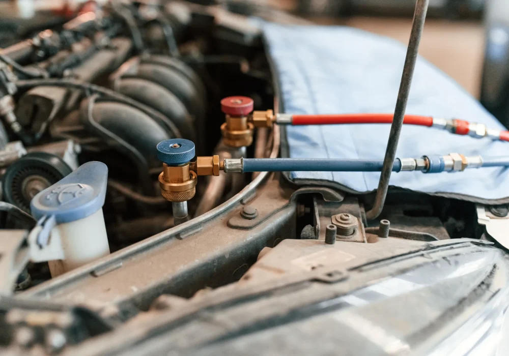 Car engine bay with blue and red hoses connected to valves for an air conditioning recharge. The hood is open, and a blue protective cover is used.