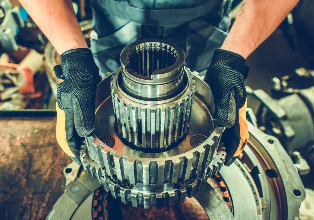A mechanic wearing gloves assembles a large metal gear on a workbench. The scene conveys precision and focus in an industrial setting.