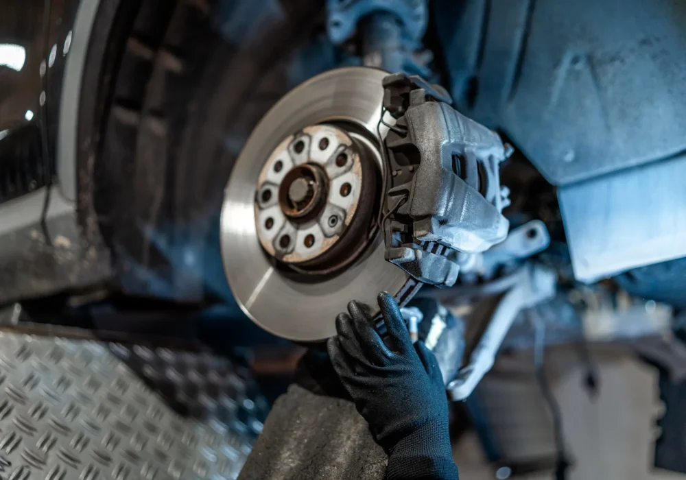 A mechanic's gloved hands inspect a car's brake disc and caliper. The setting is a workshop, suggesting maintenance or repair work.