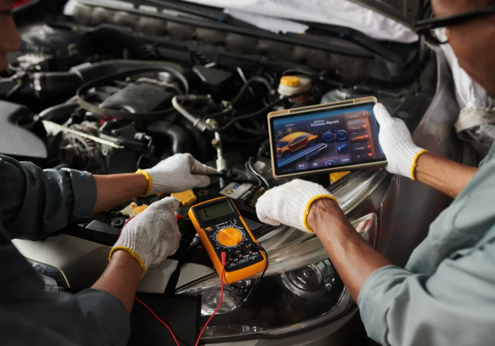 Two mechanics in gloves inspect a car engine using a digital tablet and an orange multimeter, conveying teamwork and technology in automotive repair.