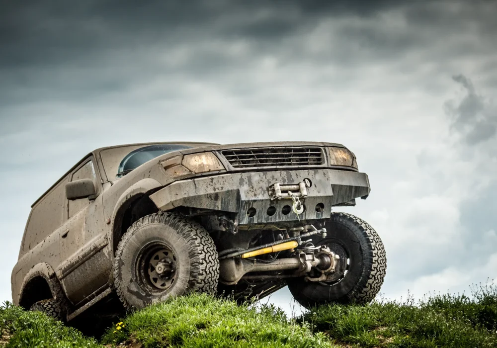 A rugged off-road SUV, covered in mud, climbs a grassy hill against a cloudy sky, conveying adventure and resilience in a challenging landscape.