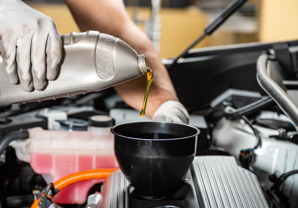 A person wearing gloves pours golden motor oil from a gray container into a funnel in a car engine bay, conveying careful maintenance and precision.